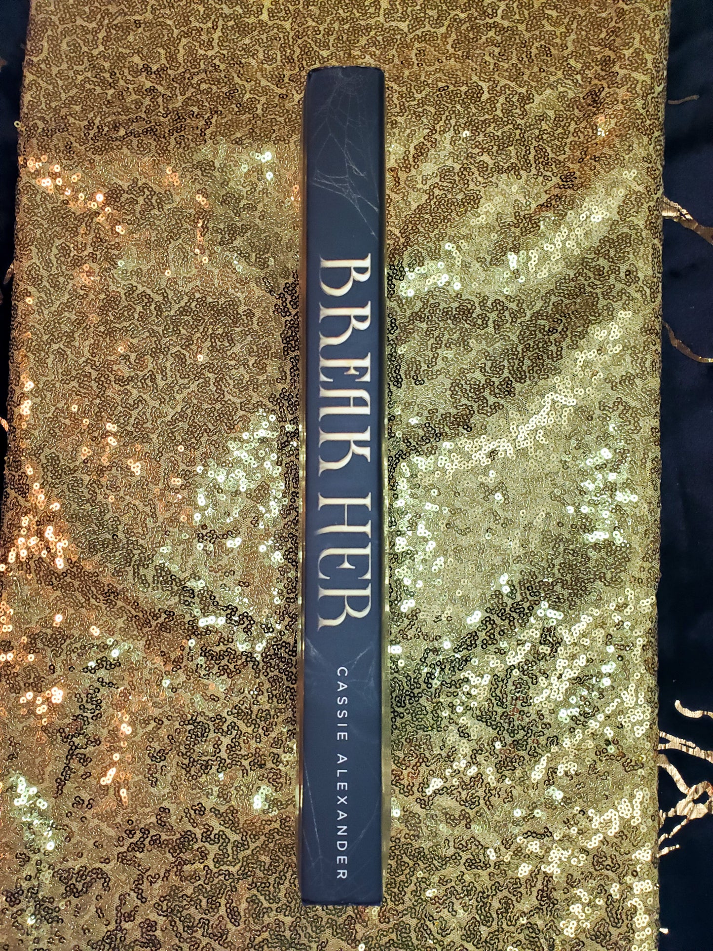 Hardcover of Break Her by Cassie Alexander on a gold sequined background. Image shows the spine.
